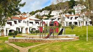 Un parque infantil con tobogán y estructuras para escalar, rodeado de un muro de piedra y un camino pavimentado.