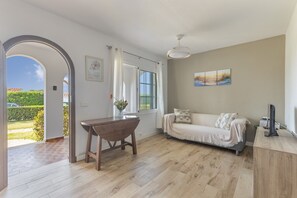 Een kamer met houten vloer, een witte bank, een tafel, een plafondventilator en een raam met gordijnen.