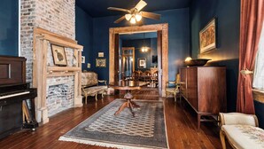 A room with a fireplace, a piano, a wooden floor, a ceiling fan, and a dining area visible through an open doorway.