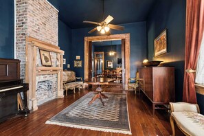 A room with a fireplace, a piano, a wooden floor, a ceiling fan, and a dining area visible through an open doorway.