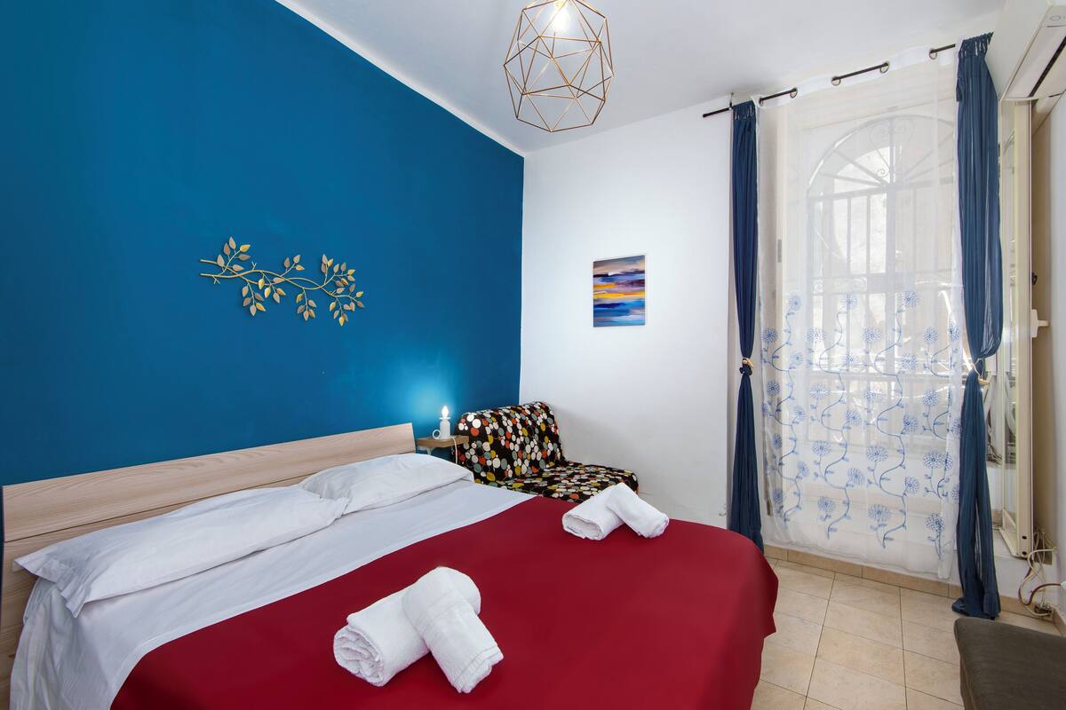Una habitación de hotel con una cama, una silla, una obra de arte en la pared y una ventana con cortinas.
