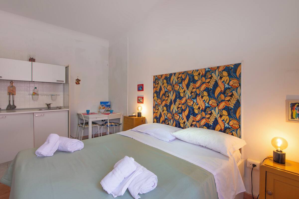 Habitación de hotel con cama, escritorio, silla, mesita pequeña, mesita de noche con lámpara y un tapiz floral en la pared.