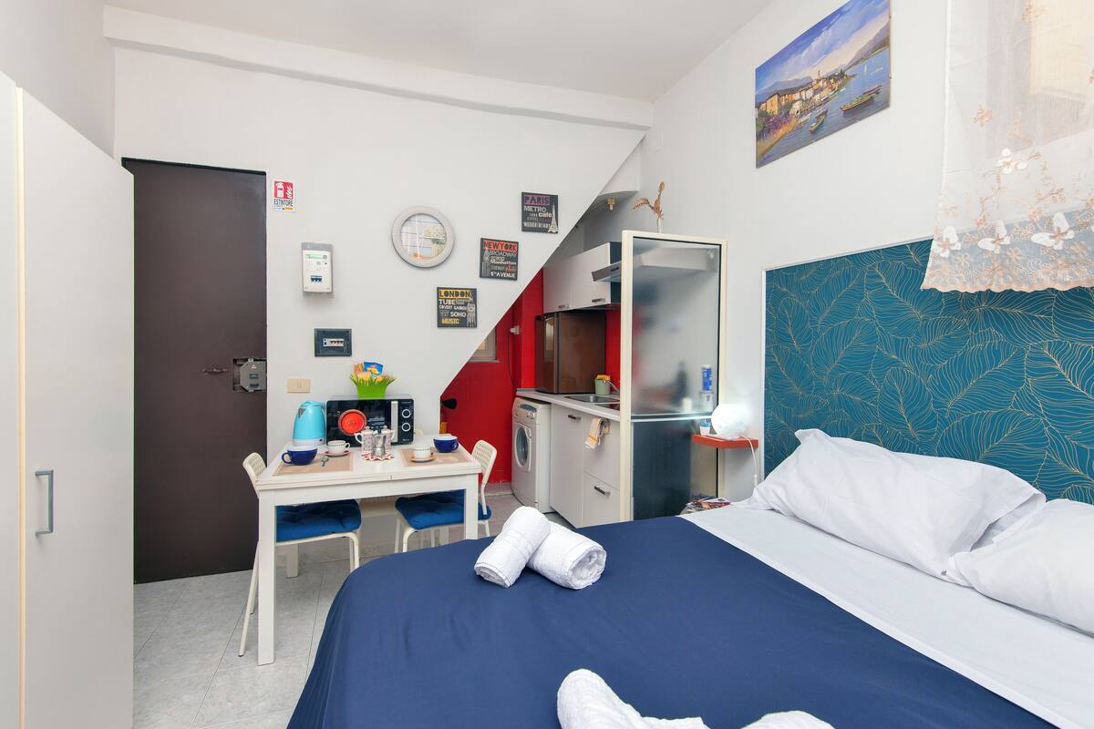 Habitación de hotel con cama, escritorio con silla, lavadora, pequeña cocina y pared roja.