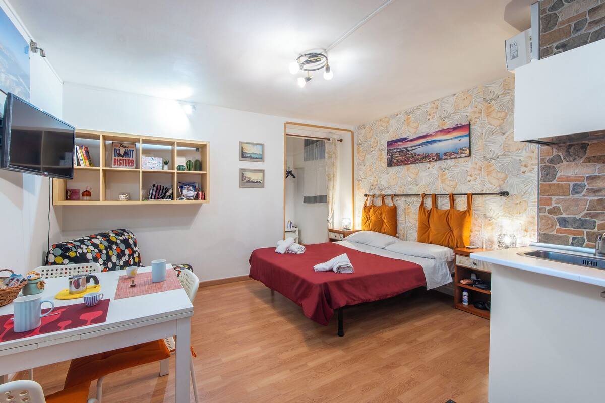 Una habitación de hotel compacta con cama, una mesita, televisor y kitchenette.
