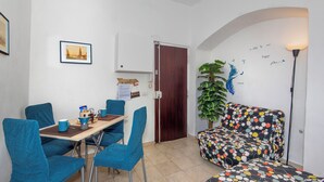 Une petite pièce comprenant une table à manger, des chaises bleues, un canapé recouvert d’un tissu à pois colorés, une plante en pot et un mur orné d’un tableau encadré et de quelques inscriptions.