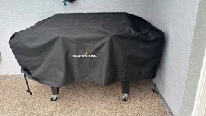 Ein Tisch im Freien mit Überdachung, mit dem BLACKSTONE Logo, vor einer strukturierten Wand mit blauer Blumendekoration.
