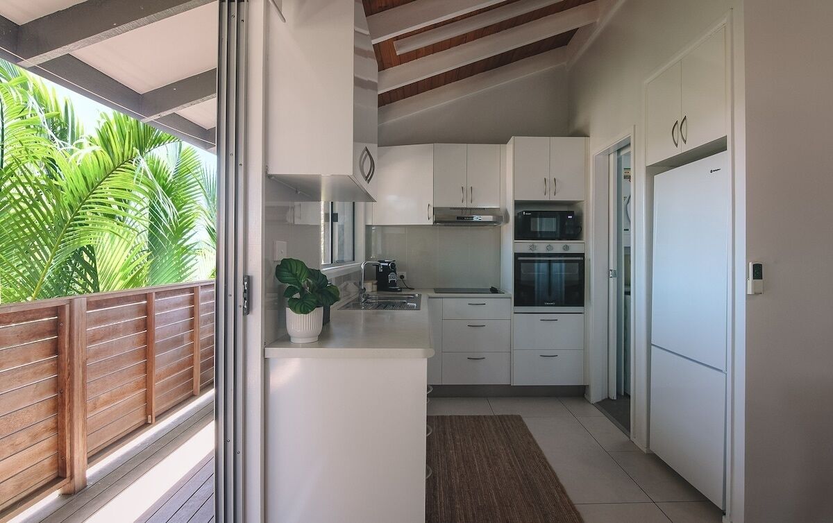 A modern kitchen with white cabinets, a built-in oven, and a large window overlooking a balcony with a wooden railing and tropical plants.