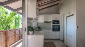 Een moderne keuken met witte kasten, een ingebouwde oven en een groot raam met uitzicht op een balkon met een houten hekwerk en tropische planten.