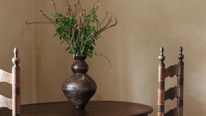 Een ronde houten tafel met een donkerbruine vaas vol groene planten en kleine witte bloemen. Er staan twee houten stoelen met zwart-wit gestreepte rugleuningen rondom de tafel.