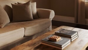 Een woonkamer met een beige bank, een houten salontafel en een raam met dunne gordijnen.