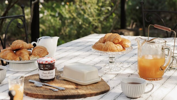 Een ontbijt buiten met croissants, boter, jam, koffie en een karaf sinaasappelsap op een tafel.