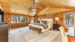 A wooden-paneled bedroom with a large bed, two bedside tables, a bench, and a view of trees through large windows. - Bua Northwoods Chalet on the Peninsula (Crystal Falls)