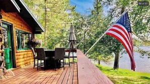 A wooden deck with a table and chairs, overlooking a lake with an American flag. - Bua Northwoods Chalet on the Peninsula (Crystal Falls)