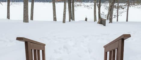 A snow-covered path leading to a wooden bench with a view of trees and a body of water in the background.
