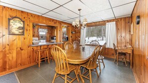 A wooden lodge dining area with a large round table, wooden chairs, a kitchen area, and a view of a snowy landscape through the windows. - Bayview Cottage 3BR Fireplace Lakefront (Saint Germain)