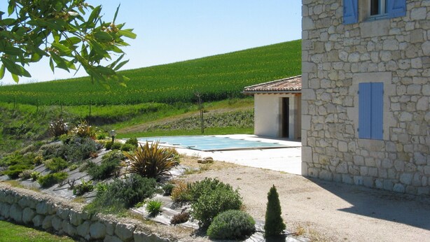 A stone house with a swimming pool, surrounded by a landscaped garden and a vineyard in the background.