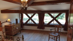A room with wooden beams, a chandelier, a bed, a wooden dresser, a chair, and large windows with curtains.