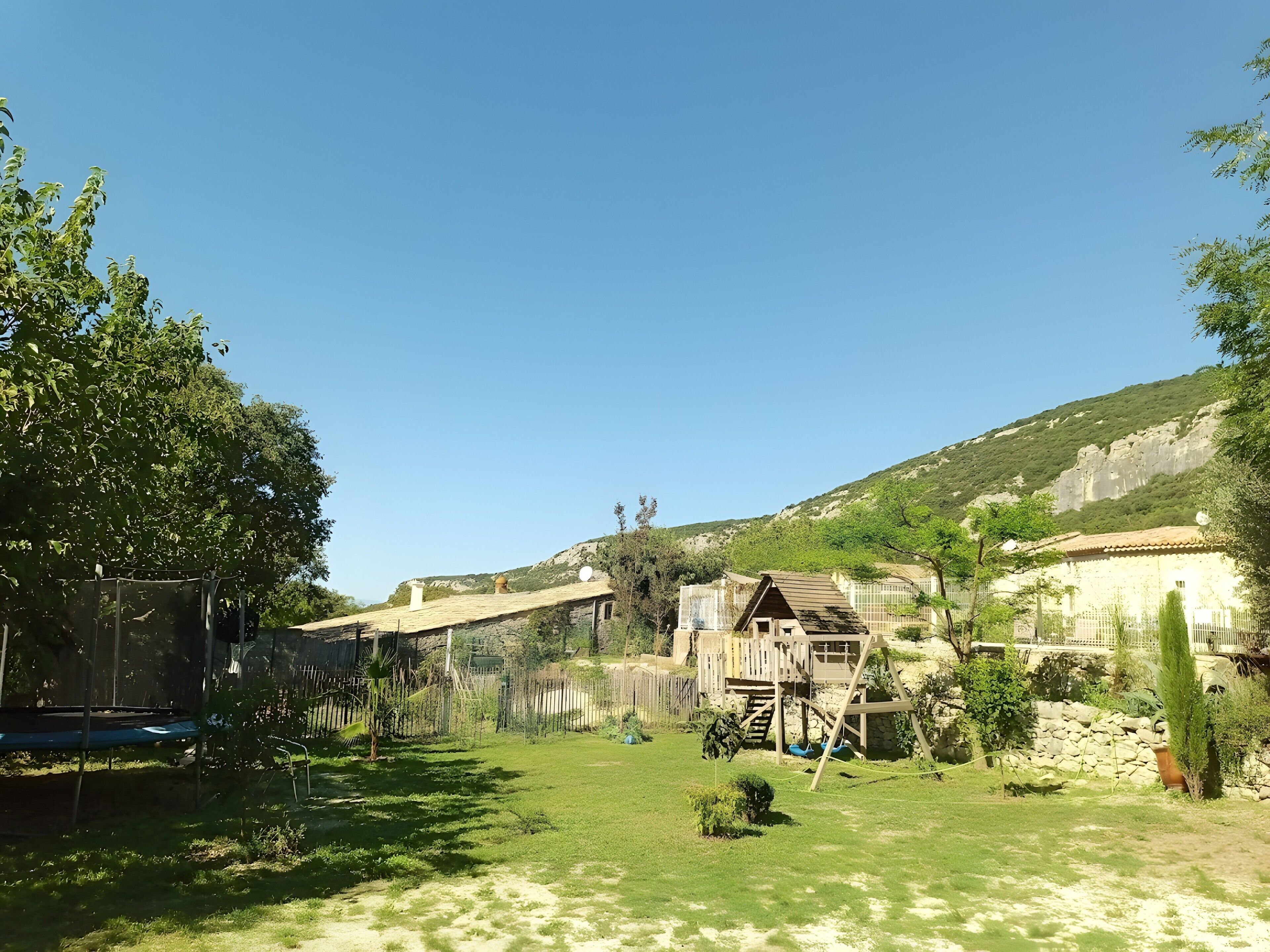 Zona residencial con jardín césped, un trampolín, estructura de juego de madera y un muro de piedra.