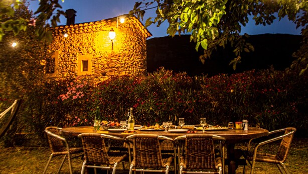 An outdoor dining area with a stone building in the background, surrounded by foliage and flowers.