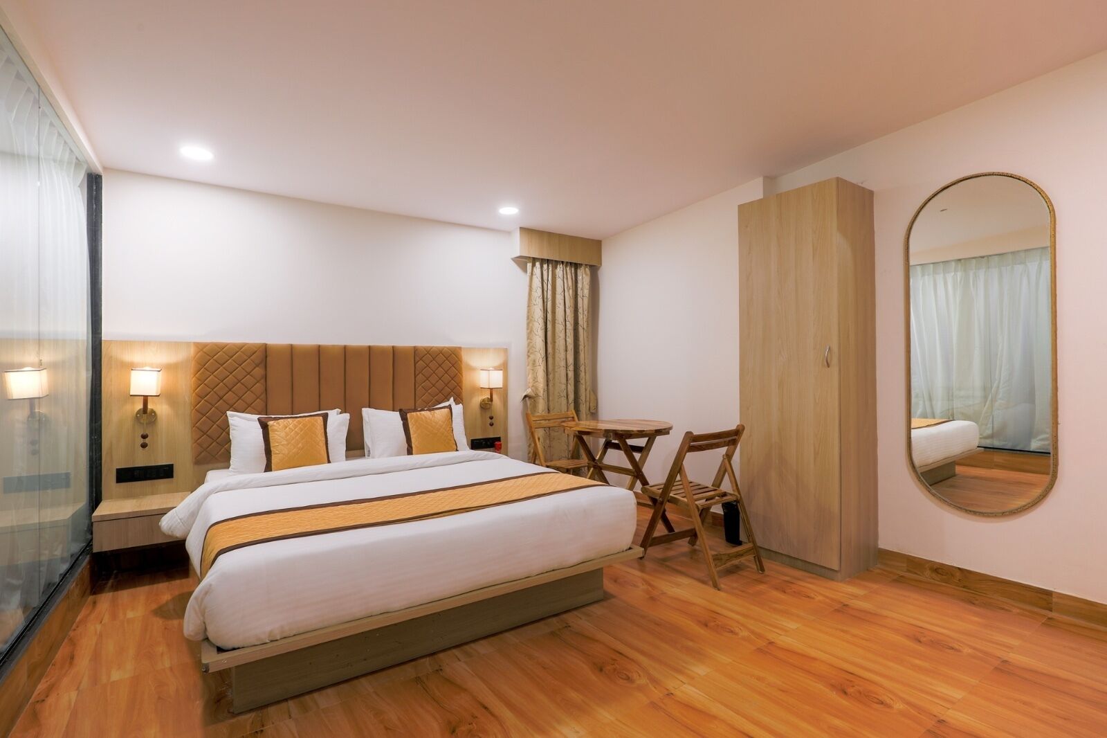 A hotel room with a large bed, two bedside tables with lamps, a wooden desk with two chairs, a wardrobe, and a mirror on the wall.