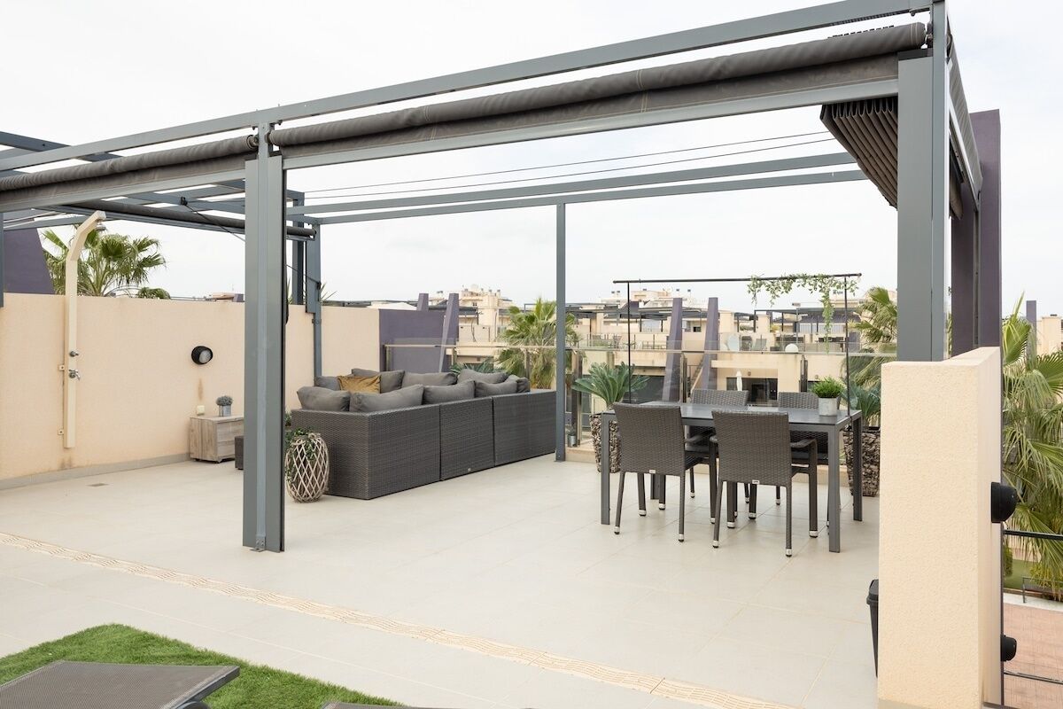 Eine überdachte Terrasse im Freien mit einer Couch und einem Esstisch, umgeben von einer Stahlkonstruktion.