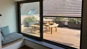 Een kamer met een grote schuifdeur die uitkomt op een terras buiten, met houten meubels en potplanten.