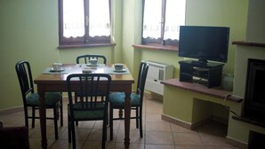 Un coin repas comprenant une table en bois, quatre chaises, un téléviseur posé sur un meuble et une fenêtre avec des rideaux.