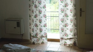 Une chambre avec un lit, un radiateur et une fenêtre donnant sur un paysage, la fenêtre étant ornée de rideaux à motifs floraux.