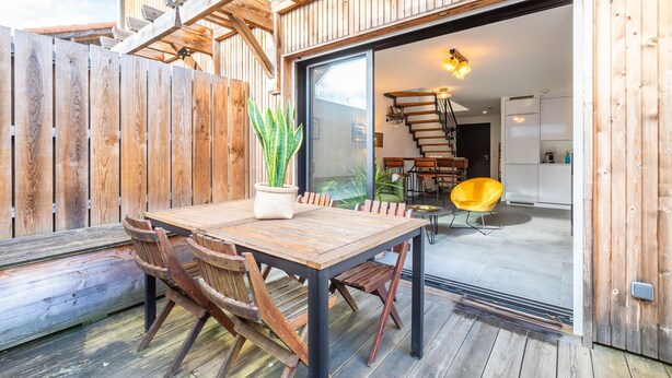 Eine Holzterrasse mit Tisch und Stühlen, einem Blumentopf und einer Treppe, die zu einem Innenbereich mit Küche und Essbereich führt.