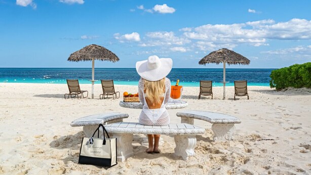 A beach scene with a woman in a white hat, a table with a drink, and chairs under thatched umbrellas.