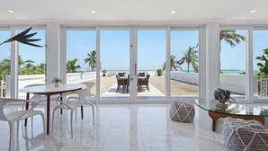A modern interior with a large glass door leading to a beach view, a dining area with white chairs, and a glass table.