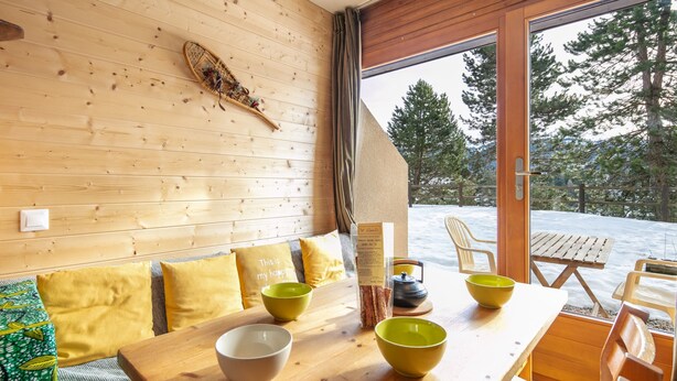Ein Holzinterieur mit einem für das Essen gedecktem Tisch, einer Bank mit gelben Kissen und Blick auf eine verschneite Landschaft durch eine Glastür.