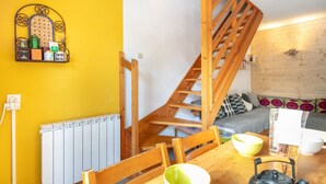 Ein kompakter Wohnraum mit einer Holztreppe, einem Esstisch mit Stühlen, einem Wasserkocher und einem Regal mit diversen Gegenständen.