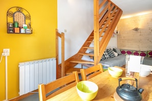 Un espacio habitable pequeño con una escalera de madera, una mesa de comedor con sillas, un hervidor y un estante con diversos objetos.
