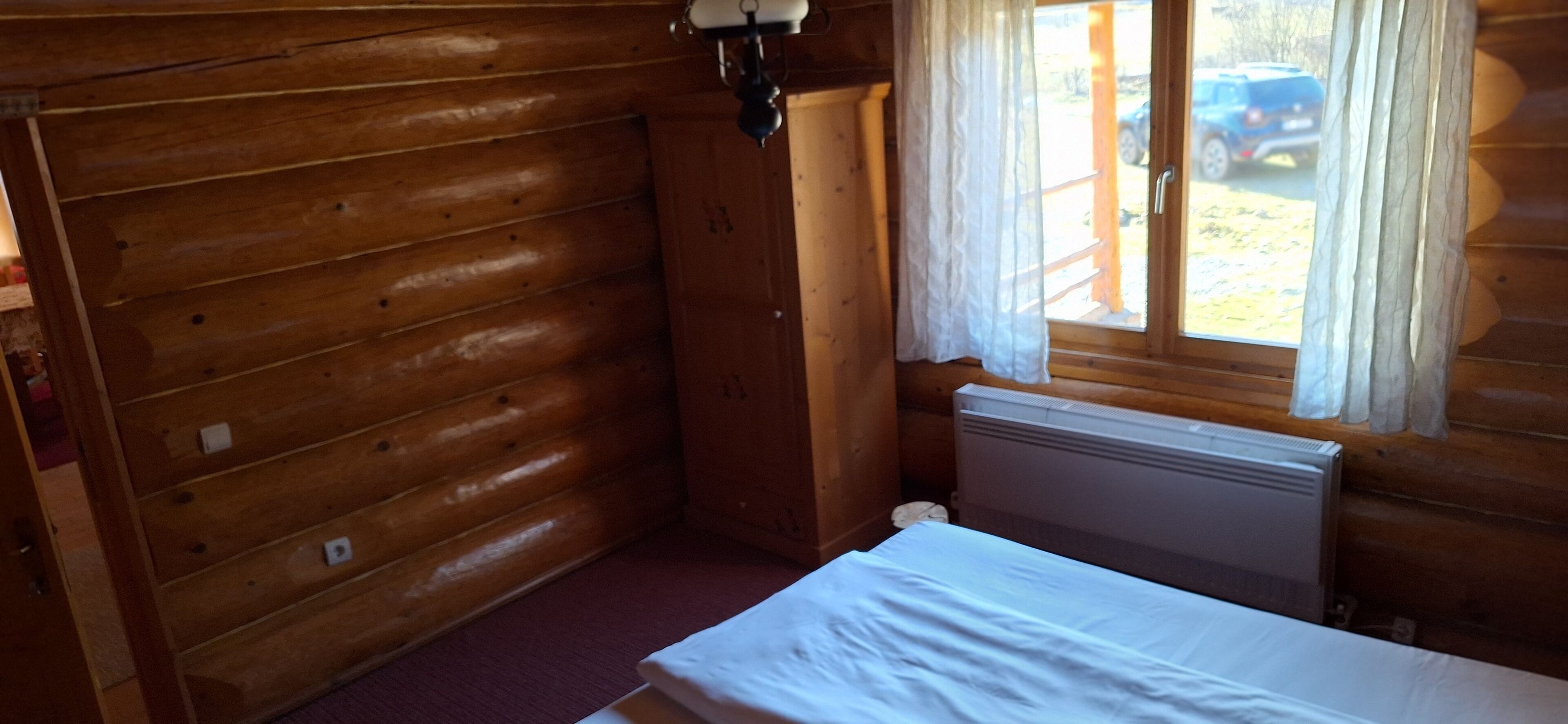 A wooden cabin room with a bed, a window with curtains, and a radiator.