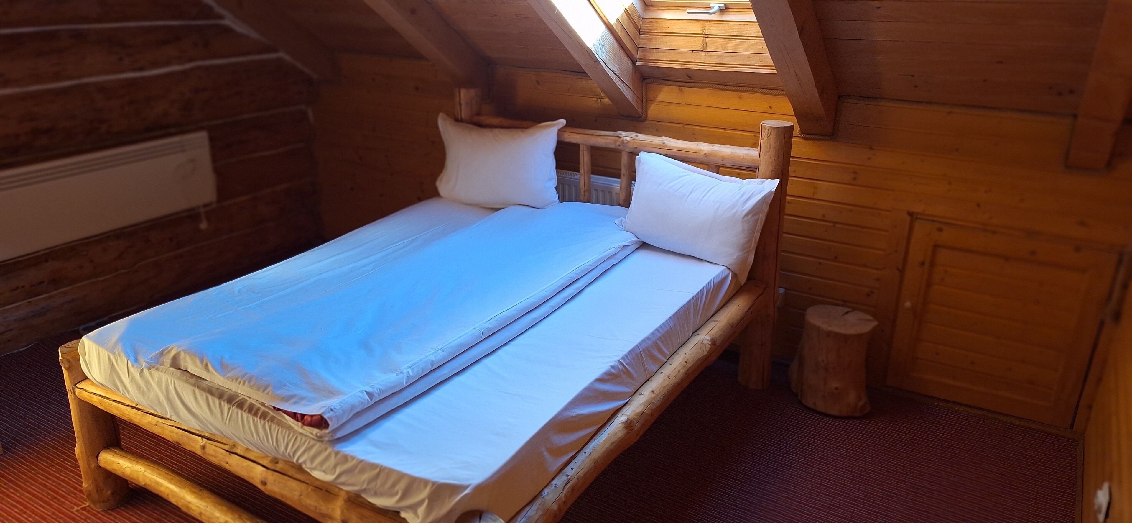 A wooden bed in a cabin with a sloped ceiling and a small wooden stool.