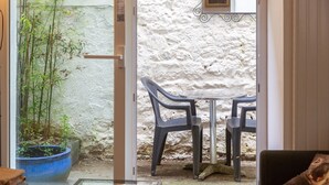 Una habitación con una puerta de cristal que da a un espacio exterior con una pared blanca, una maceta azul y dos sillas de plástico.
