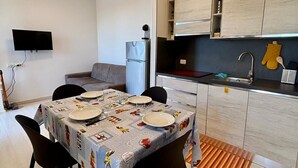 Moderni keittiö, jossa on integroidut kaapit, neljän hengen ruokapöytä, seinälle asennettu taulutelevisio ja jääkaappi.