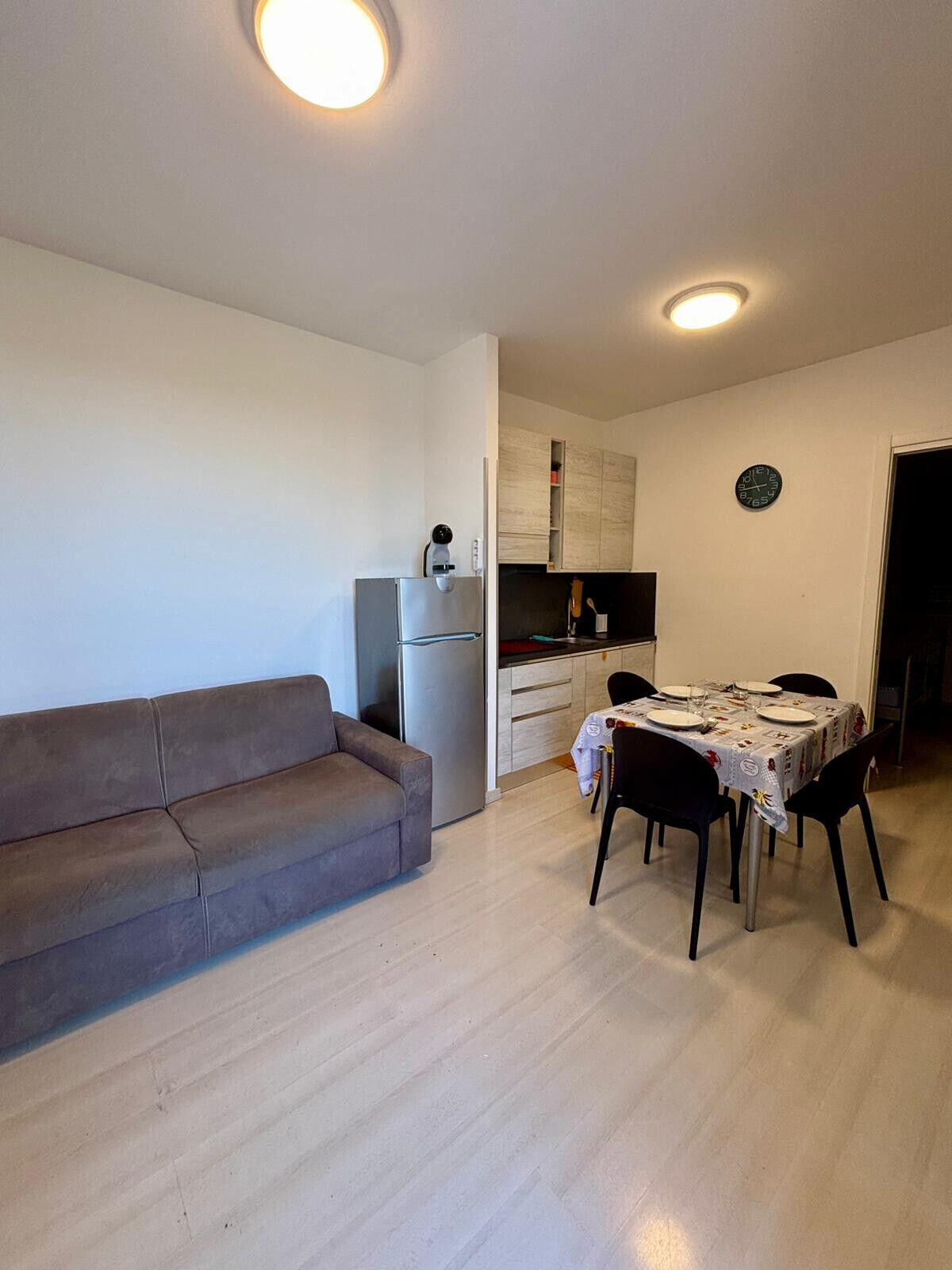 Una sala moderna con un sofá, una mesa de comedor para cuatro personas, una zona de cocina con armarios y un reloj en la pared.