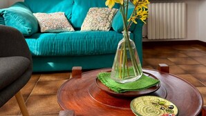 A teal sofa with patterned cushions, a clear vase with yellow flowers, a wooden coffee table with a green placemat, and a framed painting on the wall. - Olinda House in Lazise (Lazise)
