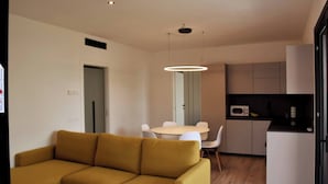 Un soggiorno moderno con un divano giallo, un tavolo da pranzo con sedie, un angolo cottura con pensili e un forno a microonde, e una lampada a sospensione sopra il tavolo.