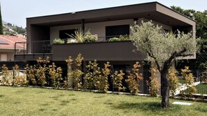 Un edificio moderno a due piani con tetto piatto, un balcone al piano superiore e un piccolo giardino con diverse piante e un albero.