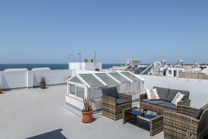 Eine Dachterrasse mit Korbmöbeln, einem Glashaus und Blick auf das Meer.