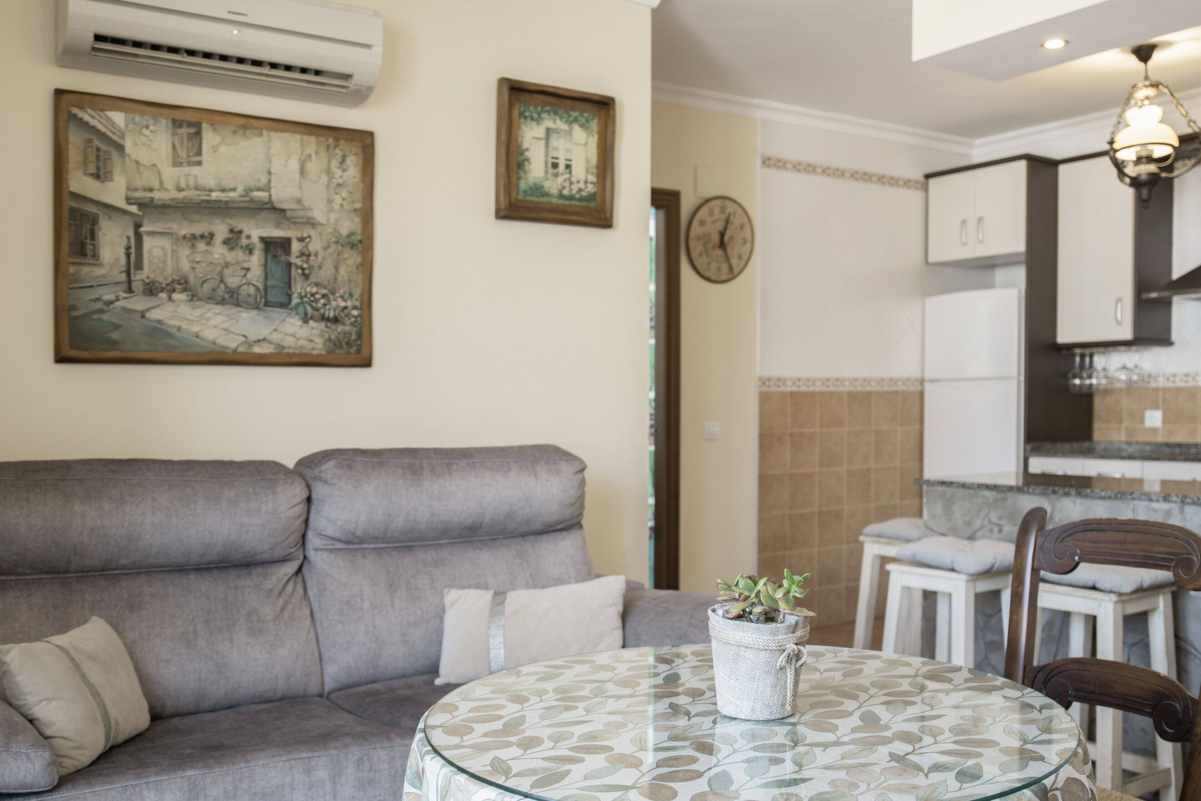 A living room with a grey sofa, a round glass table, a clock, and a kitchen area in the background.