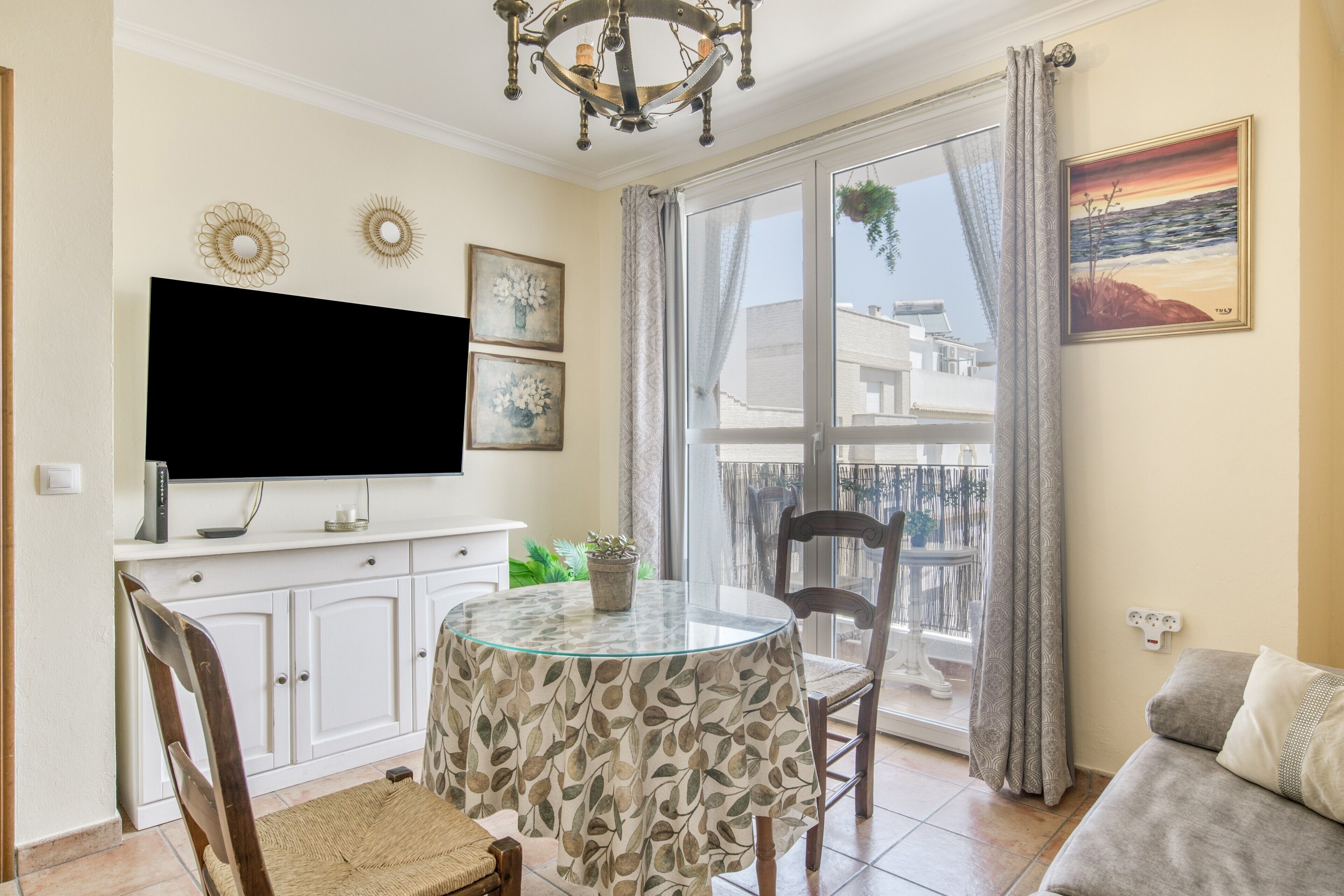 A dining area with a glass table, chairs, a television, and a balcony with curtains.
