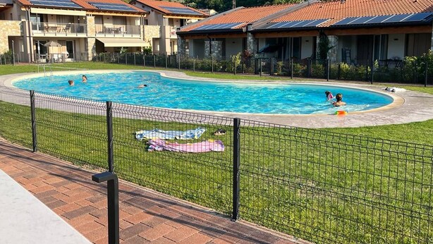 En swimmingpool omgiven av ett inhägnat område med en gräsyta, en gångstig av tegelstenar och byggnader med röda tegeltak i bakgrunden.