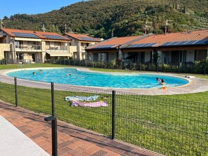 A swimming pool surrounded by a fenced area with a grassy lawn, a brick pathway, and buildings with red-tiled roofs in the background. - Villetta Sabri - Vacation Home in Garda (Garda)