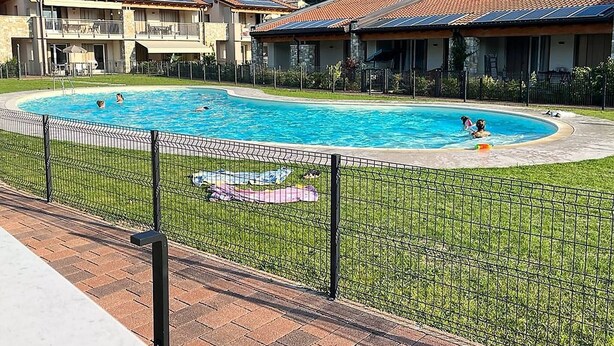 A swimming pool surrounded by a fenced area with a grassy lawn, a brick pathway, and buildings with red-tiled roofs in the background. - Villetta Pima - Holiday Home in Garda (Garda)