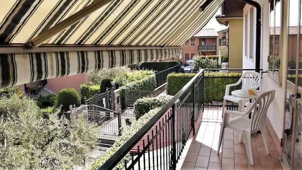 Un balcón con toldo a rayas, una pequeña mesa con sillas y vistas a una zona residencial con casas y vegetación.