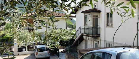 A residential area with a white building, a staircase leading to a balcony, and two parked cars.